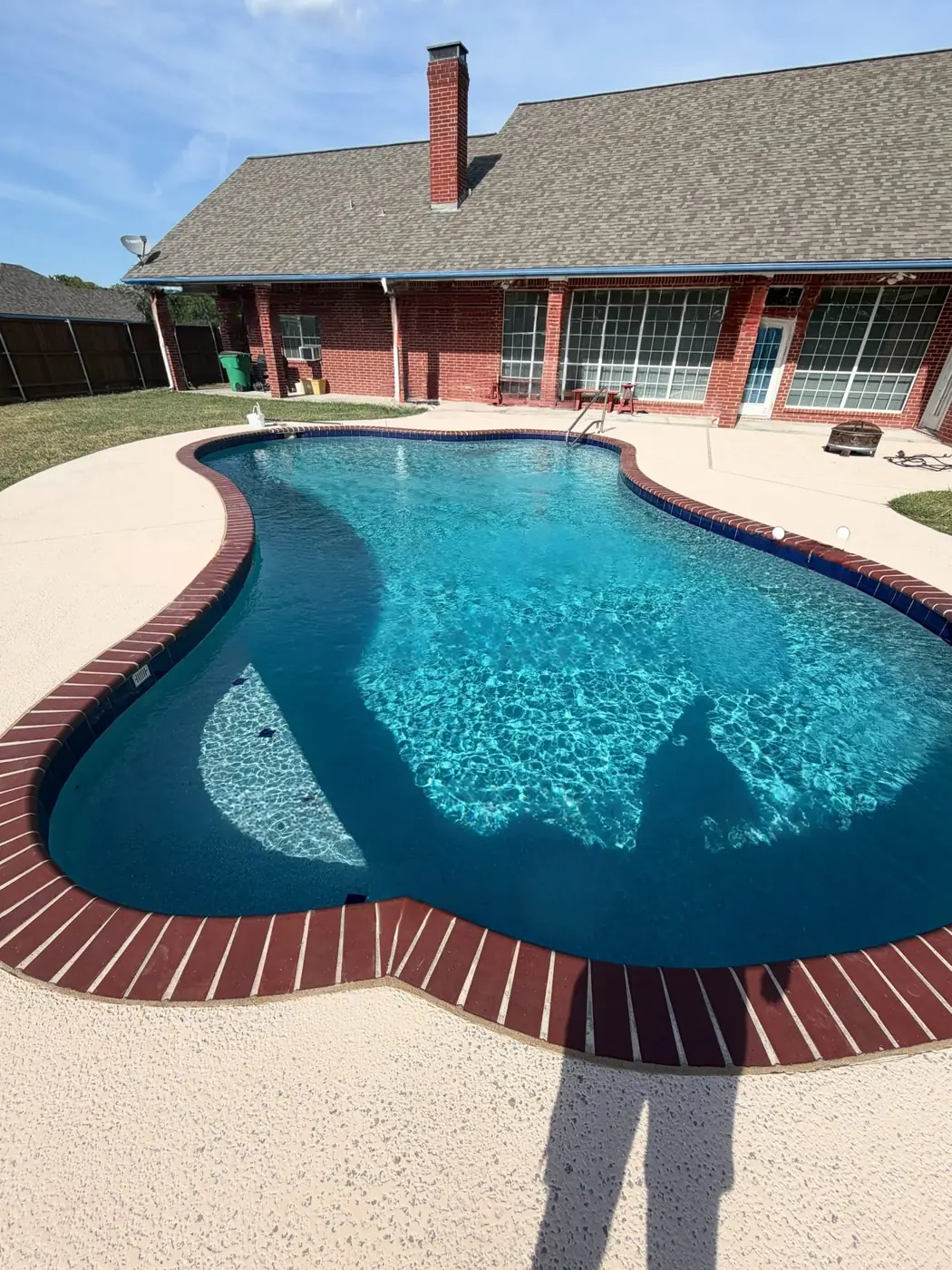 A freeform backyard pool with red brick coping beside a brick home in Greenville, Texas